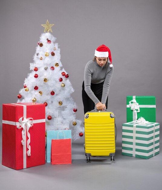 Woman wearing Santa Claus hat with luggage next to Christmas tree