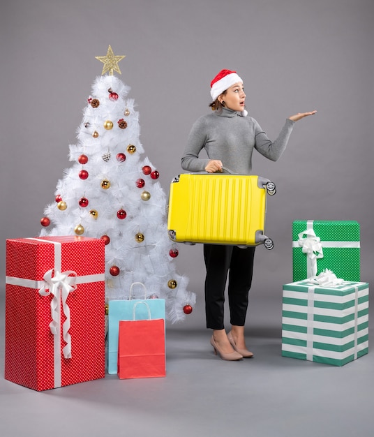 Woman wearing Santa Claus hat with luggage next to Christmas tree