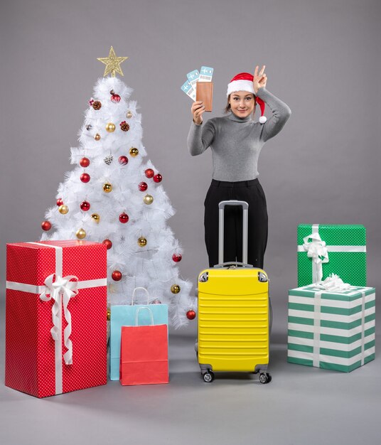 Woman wearing Santa Claus hat with luggage next to Christmas tree