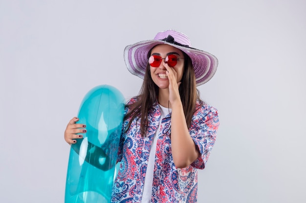 woman wearing summer hat and red sunglasses holding inflatable ring shouting or calling someone with hand near mouth positive and happy standing on white