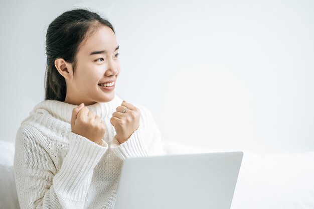 A woman wearing a white shirt on the bed and playing laptop happily.