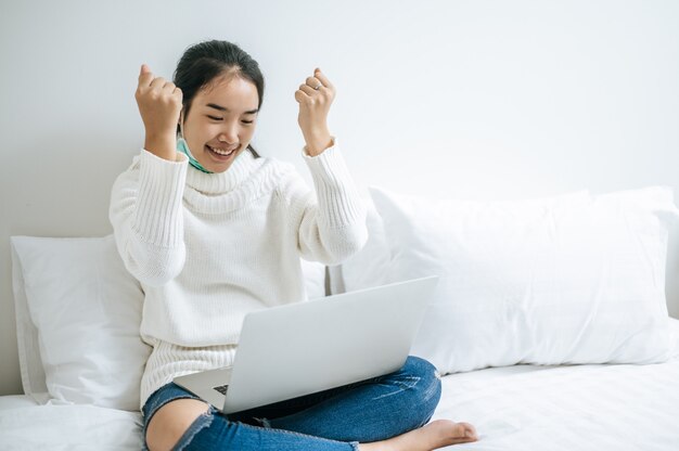 A woman wearing a white shirt on the bed and playing laptop happily.