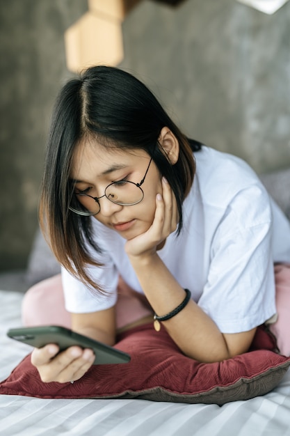 Free photo woman wearing a white shirt lying on bed and playing smartphone.