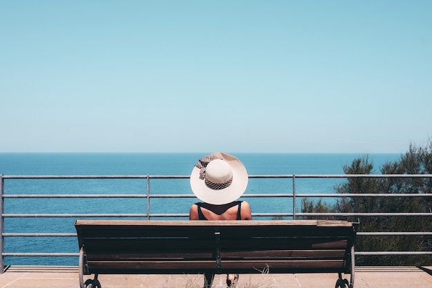 Free photo woman wearing white straw hat sitting on bench looking at sea