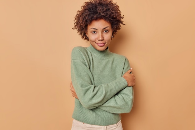 woman wears casual jumper from soft fabric looks tenderly at camera has healthy skin poses against beige studio. Beautiful female model enjoys good atmosphere.