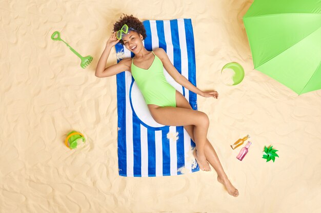 woman wears green bikini and snorkeling glasses lies on blue striped towel enjoys summer vacation spends free time at sandy beach sunbathes during sunny day. Holidays.