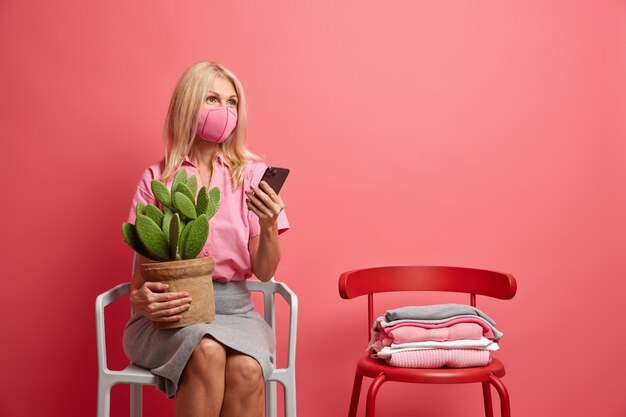 여자는 감염을 방지하기 위해 위생 마스크를 착용 코로나 바이러스는 채팅을 위해 스마트 폰을 사용하여 분홍색에 고립 된 의자에 선인장 포즈 냄비를 보유하고 있습니다.