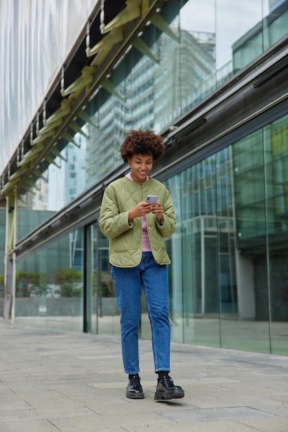 woman wears jacket and jeans chats in social networks shares files with followers smiles positively connected to wireless internet walks near modern glass shopping centre