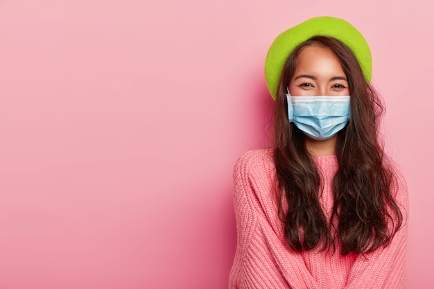 Женщина носит медицинскую маску для защиты от заболевания, зеленый берет и большой розовый свитер.