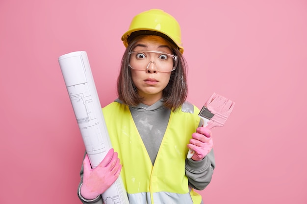 woman wears protective helmet glasses and gloves going to paint something busy with house repairing holds blueprint 