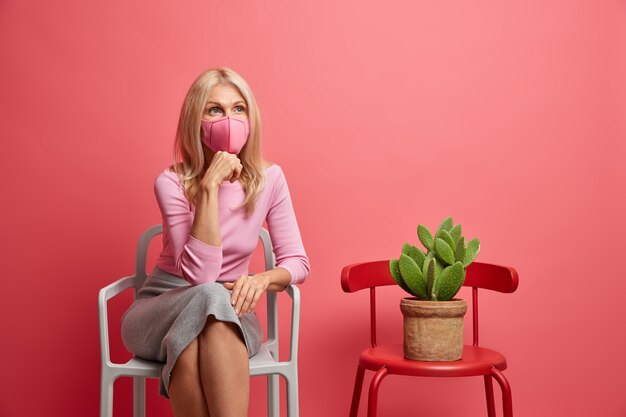 woman wears protective mask stays at home during quarantine prevents coronavirus dressed in casual jumper and skir sits alone on chair