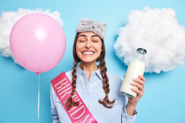 woman wears sleepmask on head holds glass bottle of milk and balloon enjoys cozy domestic atmosphere models against blue 