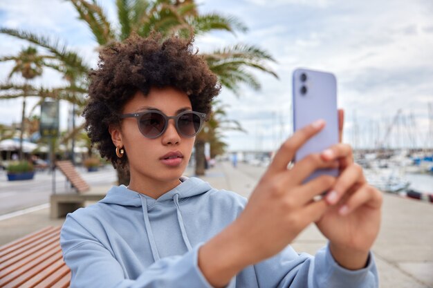 woman wears sunglasses and blue hoodie makes selfie on smartphone camera poses near sea port poses outside makes online call uses free internet