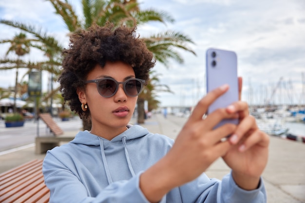 女性はサングラスをかけ、青いパーカーはスマートフォンで自分撮りをしますカメラは外の港のポーズの近くでポーズをとりますオンライン通話は無料のインターネットを使用します