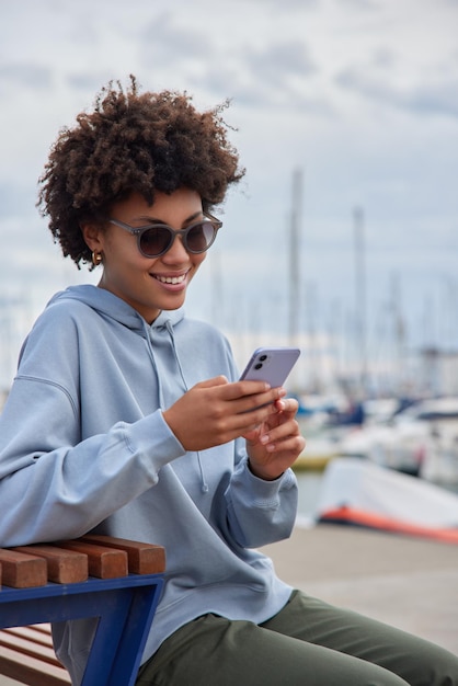 woman wears sunglasses and casual clothes uses mobile phone for chatting online rests in harbor has vacation at sea poses at harbor has cheerful expression. Lifestyle concept