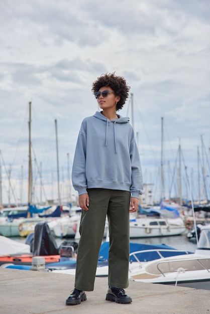 woman wears sunglasses hoodie and trousers poses on yacht harbor enjoys leisue time admires beautiful views has promenade during pastime