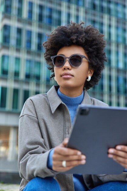 woman wears trendy sunglasses and jacket holds modern digital tablet watches tutorial video poses against modern city building uses technology for online communication