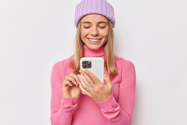 woman wears warm knitted hat and turtleneck uses mobile phone shares impressions after holidays on her blog isolated on white chats online.