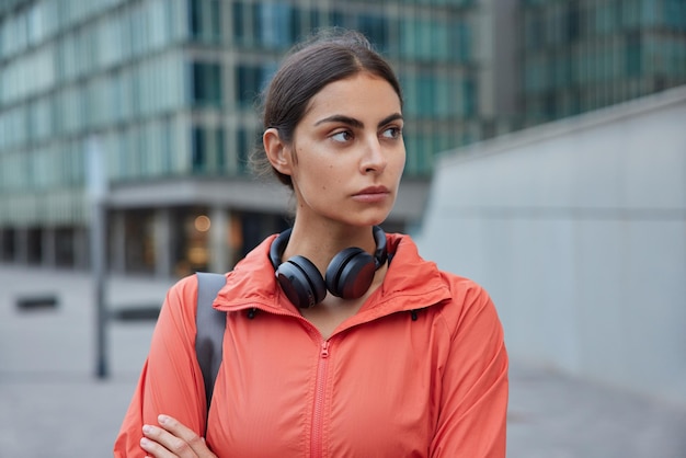 Woman wears windbreaker wears stereo headphones around neck returns fromm fitness training poses outdoors outdoor