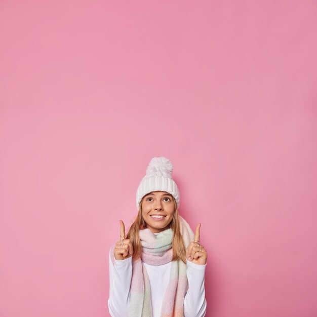 여자는 겨울 모자를 쓰고 목에 스카프를 두르고 빈 카피 공간에서 위의 분홍색 점에 대해 포즈를 취합니다.
