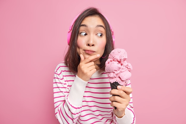 woman wears wireless headphones listens music holds chin eats delicious appetizing ice cream of pleasant flavor dressed in casual striped jumper 