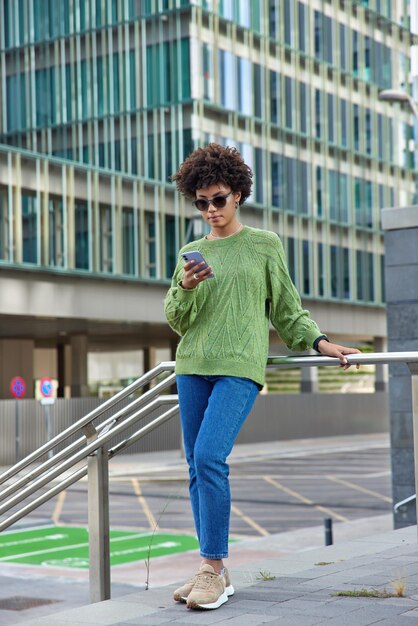女性wearstrendyサングラスカジュアルジャンパージーンズとスニーカーは携帯電話での呼び出しを待つソーシャルネットワークにメディアをアップロードする屋外で休むテキスト出版物を閲覧する