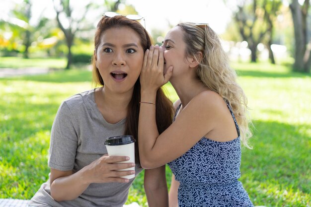 Woman whispering secret to surprised female friend in park