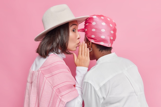  woman whispers gossips in friends ear weas hat and shirt isolated on pink 