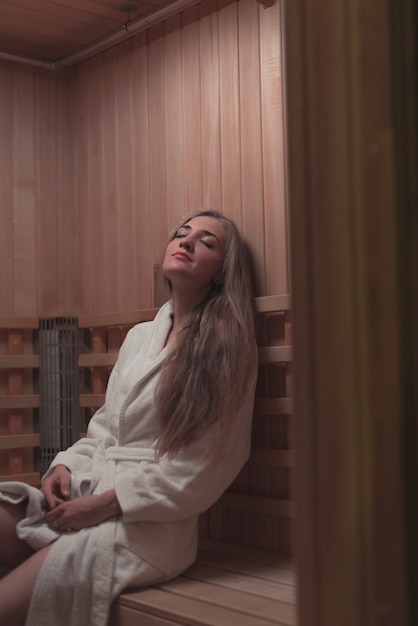 Donna in accappatoio bianco che si siede sul banco di legno rilassante nella sauna