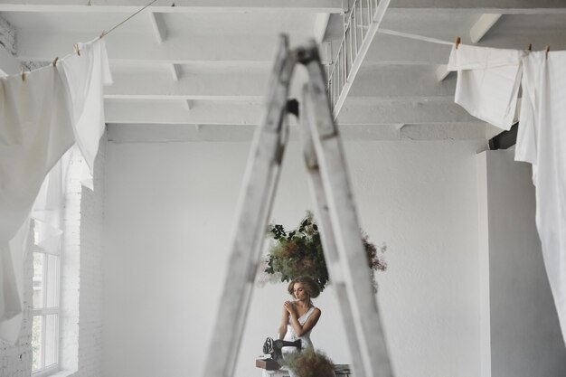 Woman in white clothes sits in a room with flowers and sewing machine