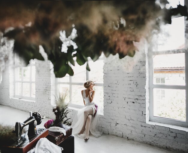 Woman in white clothes sits on the windowsill in a room with flowers and sewing machine