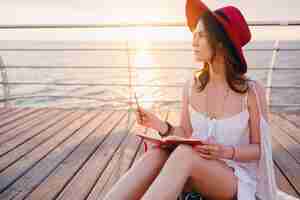 Foto gratuita donna in abito bianco seduto in riva al mare all'alba pensando e prendendo appunti nel diario in umore romantico indossando il cappello rosso