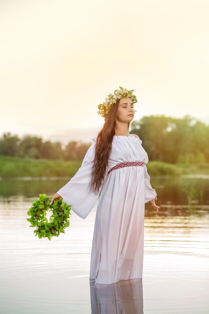 水の中の白いドレスを着た女性。川で彼女の頭に花輪を捧げるアートウーマン。湖の濡れた魔女少女、神秘的な謎の女。彼女の頭に花輪、スラブの伝統と異教