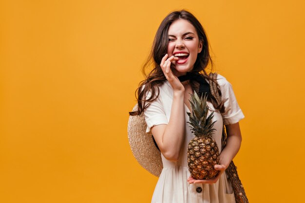 Woman in white dress with hat on her back bites her finger and holds pineapple on orange background.