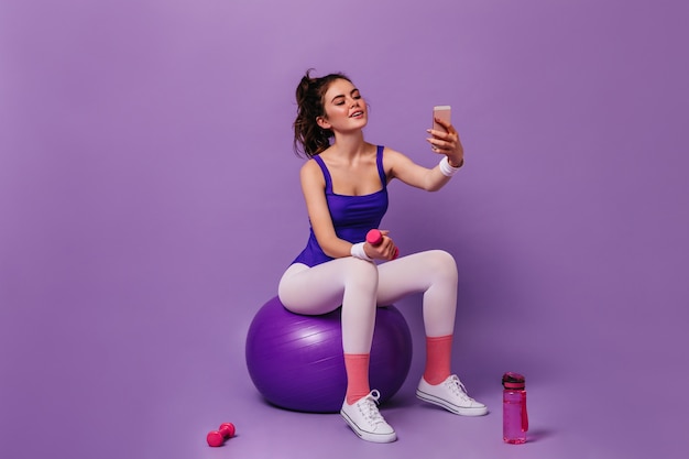 Woman in white leggings and purple bodysuit holds pink dumbbell and takes selfie on isolated wall