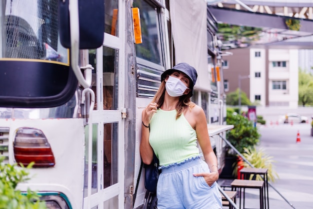 白い医療マスクの女性が街を歩く街の広場のバスカフェのそばに立つ