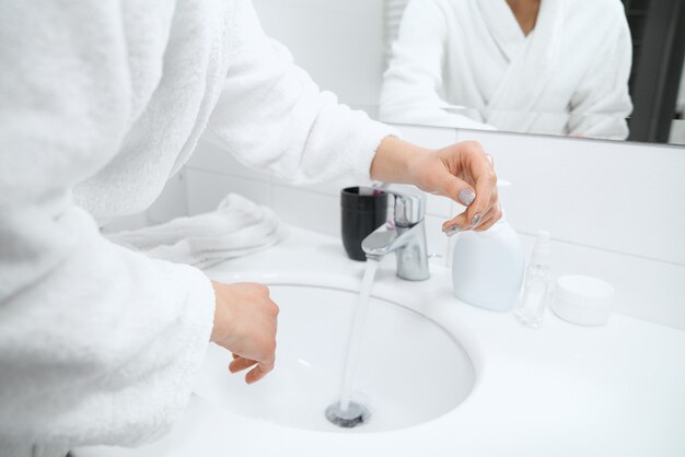 Woman in white robe standing near sink and washing hands