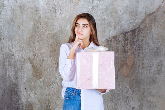 женщина в белой рубашке держит розовую подарочную коробку, обернутую белой лентой, и выглядит смущенной и нерешительной.