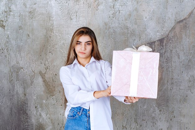 женщина в белой рубашке держит розовую подарочную коробку, обернутую белой лентой, и выглядит смущенной и нерешительной.