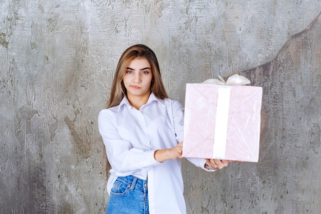 woman in white shirt holding a pink gift box wrapped with white ribbon and looks confused and hesitating.