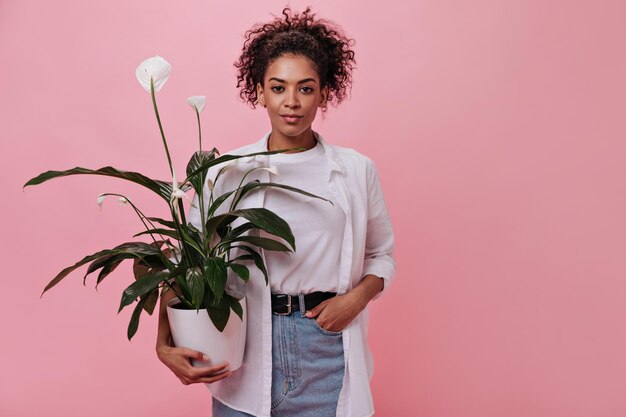 Woman in white shirt is holding flower on pink background Curly girl in denim skirt posing with pants on isolated backdrop