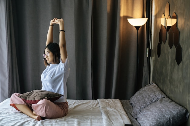 A woman in a white shirt sitting on the bed and raising both arms.