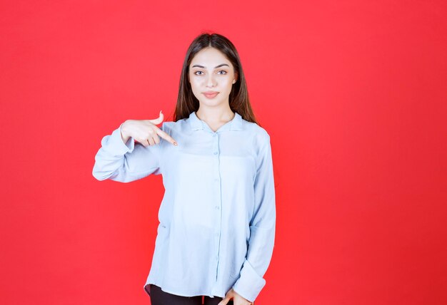 woman in white shirt standing on red wall and pointing at herself. 