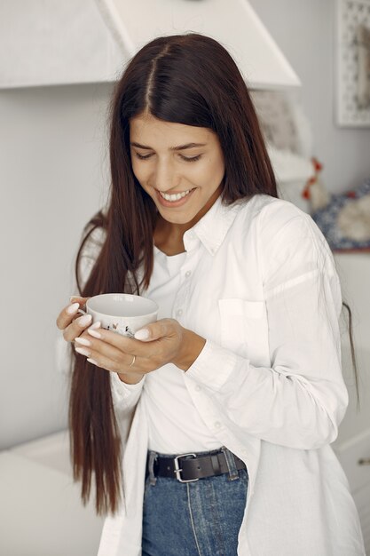 Woman in a white shirt standing in rhe kitchen and drinking a coffee