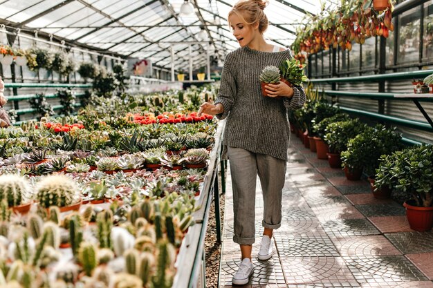 흰색 운동화를 입은 여성, 회색 헐렁한 옷차림이 식물 가게를 산책하고 선인장을 손에 쥐고 있습니다.