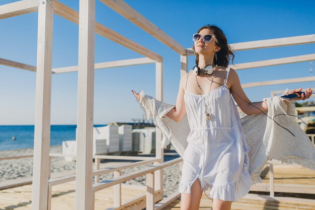 woman in white summer dress listening to music on headphones dancing and having fun