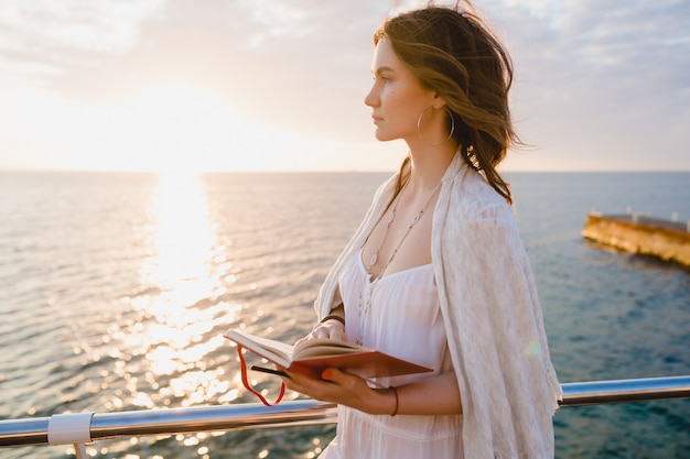 woman in white summer dress walking by the sea on sunrise with diary book in romantic mood thinking and making notes