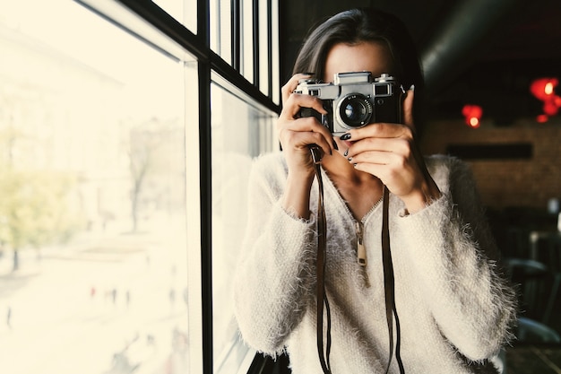 Woman in white sweater stands with camera before a bright window