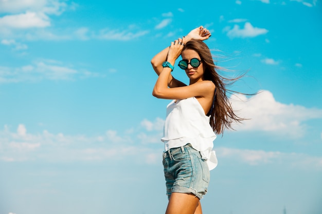 Woman in white t shirt and stylish sunglasses posing on the beach. Blue sky.