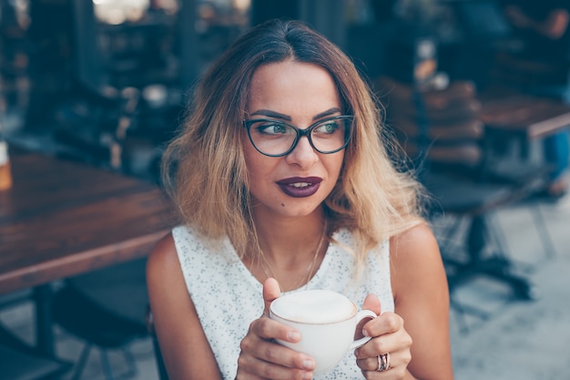 Donna in camicia bianca con texture seduto e tenendo il caffè nella terrazza del caffè durante il giorno